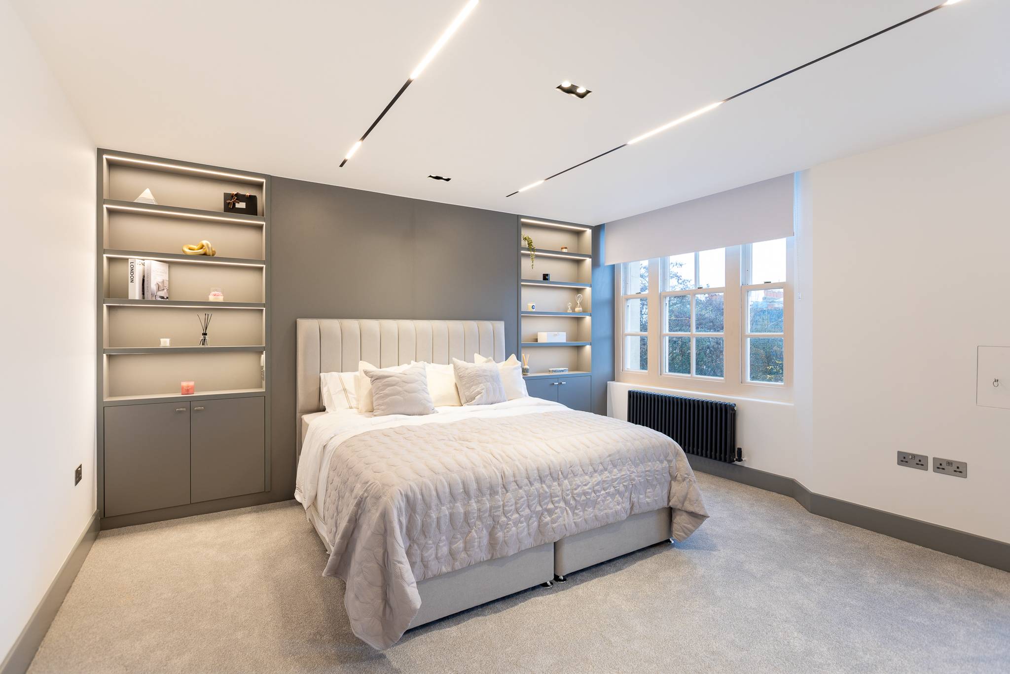 Bedroom after, with a grey panelled bed wall, lit shelving either side and recessed ceiling lights