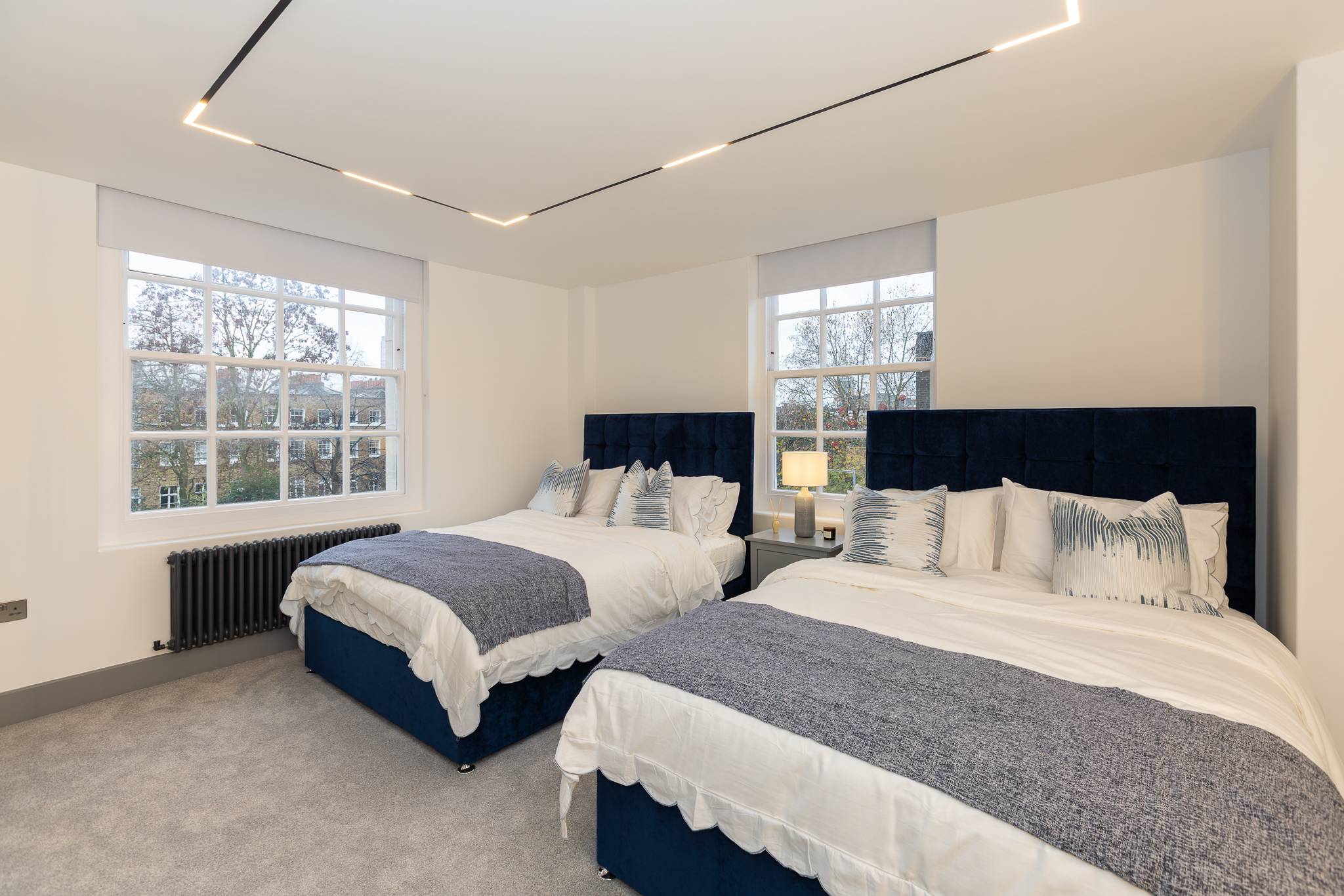 The same room after, as a twin bedroom with two navy velvet beds and linear ceiling lights